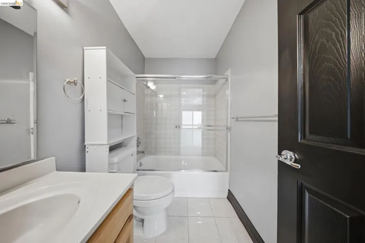Bathroom featuring a wood-finish vanity with an integrated sink, shower-over-tub with sliding glass doors, white tiled walls, white tile flooring, and an over-toilet storage unit