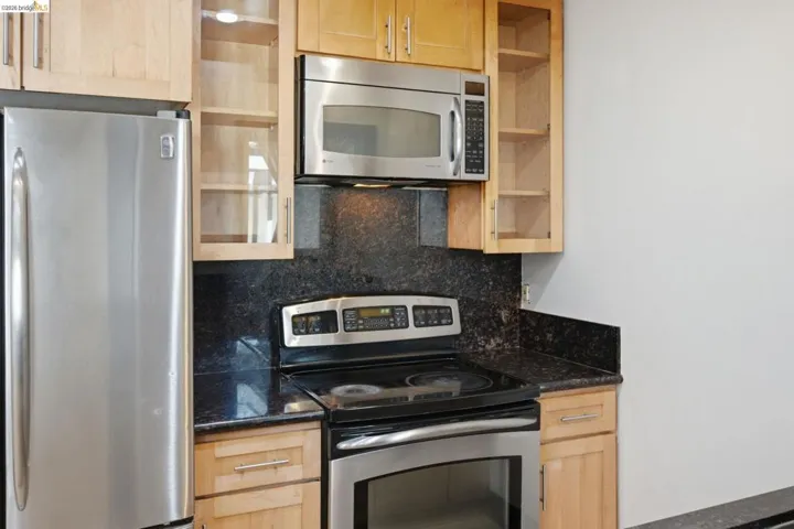 Kitchen featuring light wood-finish cabinetry, dark stone countertops, stainless steel appliances, and a matching stone backsplash