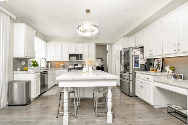 Spacious kitchen featuring an elongated island with a marble-finish countertop, white cabinetry, integrated stainless steel appliances, a gray subway tile backsplash, and light wood-finish flooring