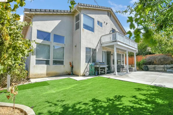 Rear exterior featuring an upper-level balcony, a lower patio area, and an artificial turf lawn