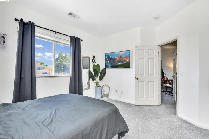 Carpeted bedroom featuring a large window, white paneled door, ceiling vent, and a mounted display