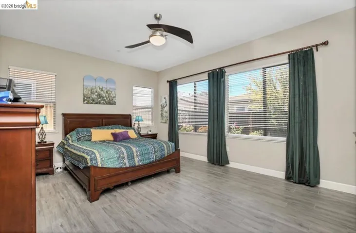 Spacious room featuring wood-finish flooring, multiple windows with blinds, a ceiling fan with integrated lighting, and neutral wall tones