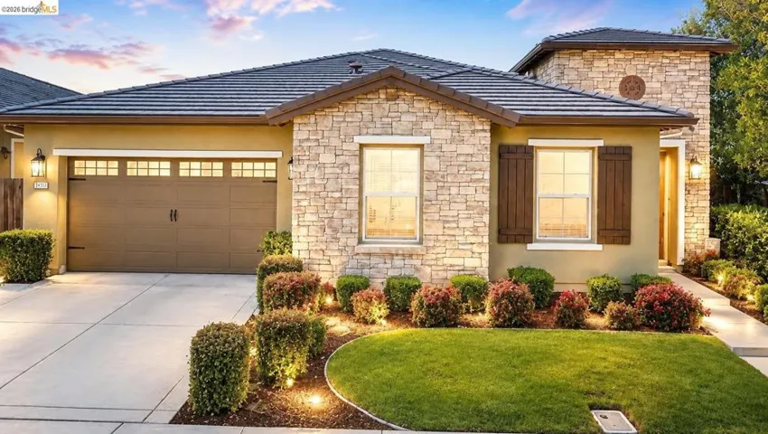 Stone facade residence featuring a two-car garage with transom windows, dark wood shutters, a covered entry, and manicured landscaping with ground lighting