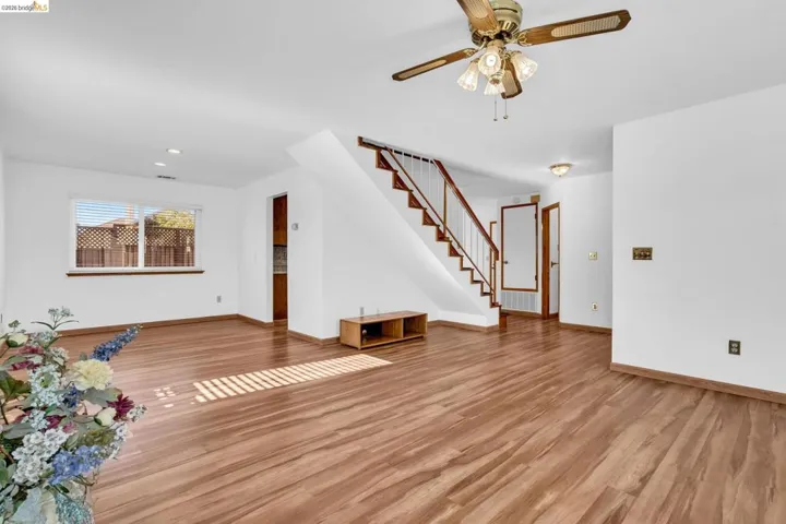 Spacious living area featuring wood-finish flooring, a ceiling fan with integrated lighting, and a staircase with wood handrails