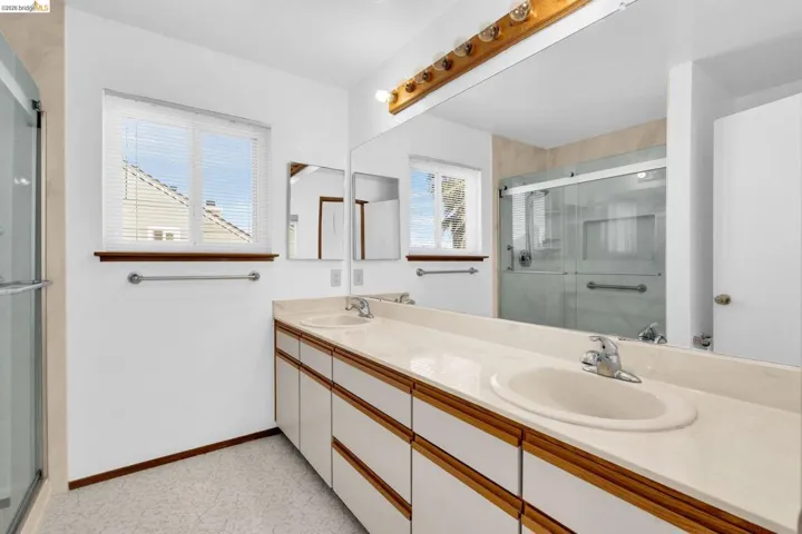 Bathroom featuring a dual vanity with integrated sinks, a wide mirror, a glass-enclosed shower, and a large window with privacy blinds