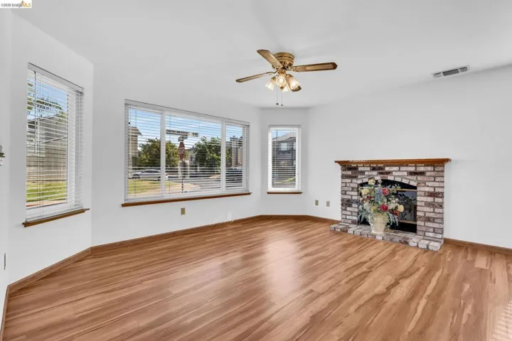 Spacious interior featuring wood-finish flooring, a brick fireplace with wood mantle, multiple windows with blinds, and a ceiling fan with integrated lighting
