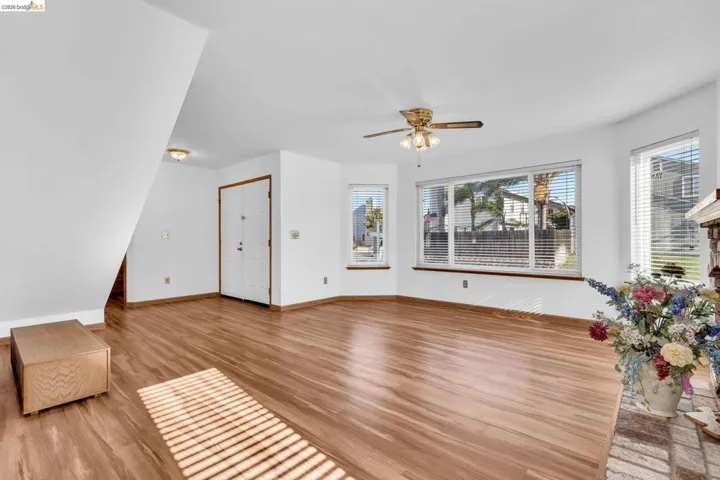 Spacious living area featuring wood-finish flooring, multiple windows with horizontal blinds, a ceiling fan with integrated lighting, and a prominent brick-faced fireplace