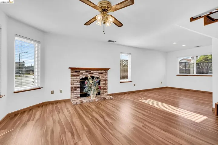 Expansive living area featuring wood-finish flooring, a brick fireplace with wood mantel, multiple large windows, and a ceiling fan with integrated lighting