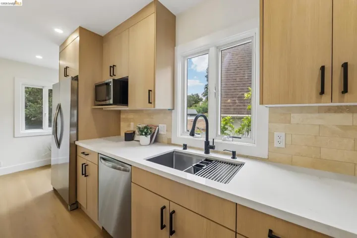 Modern kitchen featuring light wood-finish cabinetry, stainless steel appliances, white countertops, a deep undermount sink with a black faucet, and a stone-tile backsplash