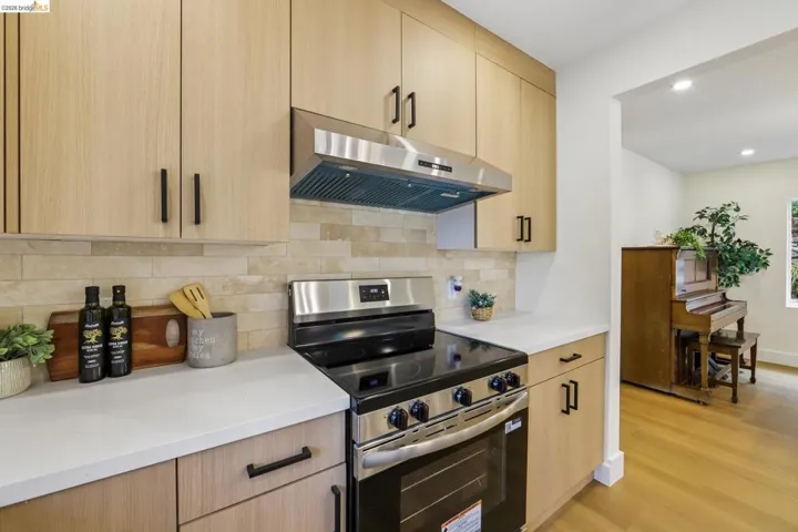 Contemporary kitchen featuring light wood-finish cabinetry, stainless steel range with matching vent hood, light-toned countertops, and a stone-style tile backsplash