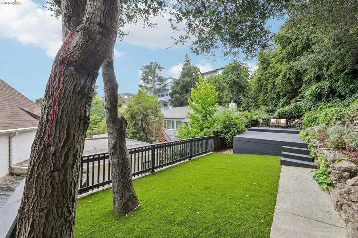 Expansive outdoor space featuring a manicured artificial turf lawn, a multi-tiered deck, a stone retaining wall, and mature trees providing natural screening