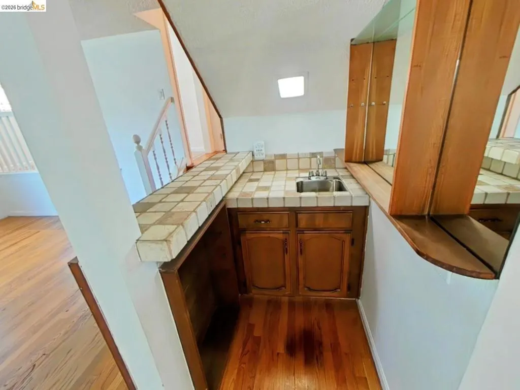 Compact wet bar featuring a tile countertop, integrated stainless steel sink, wood cabinetry, and wood-finish flooring