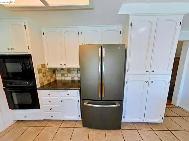 Kitchen featuring white cabinetry, a built-in black microwave and oven, a dark stainless steel refrigerator, dark countertops, and a tiled backsplash
