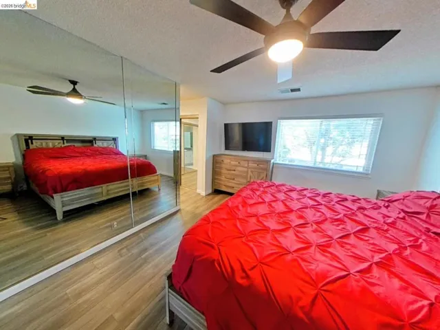 Bedroom featuring wood-finish flooring, a ceiling fan with integrated lighting, a wide window with blinds, and a mirrored wall
