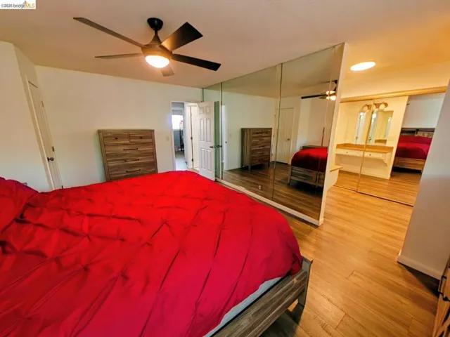 Wood-finish flooring throughout, featuring a built-in dressing vanity with overhead lighting, a modern ceiling fan with integrated lighting, and mirrored closet doors