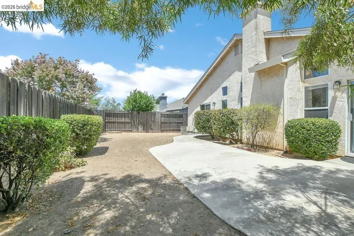 Spacious yard featuring a concrete patio, mature perimeter shrubs, a wood fence, and a stucco exterior with a prominent chimney