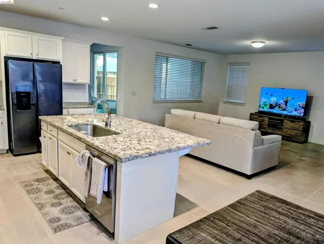 Open-concept living space featuring a kitchen island with stone countertop and integrated sink, stainless steel dishwasher, white cabinetry, and large format tile flooring
