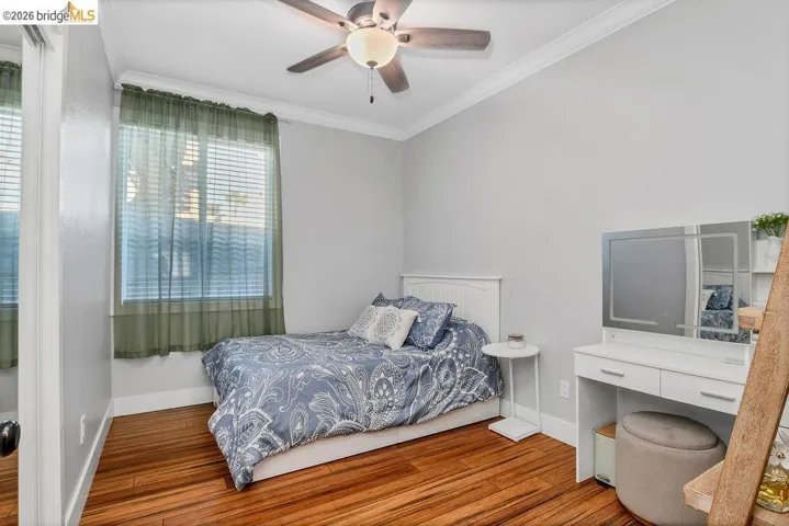 Wood-finish flooring anchors this room, complemented by a window with blinds, crown molding, and a ceiling fan with integrated lighting