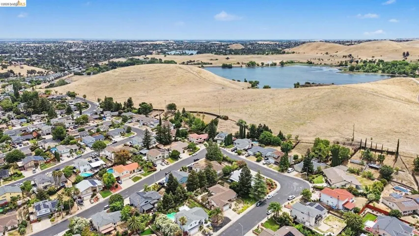 Residential area featuring paved streets and varied rooflines, bordered by expansive natural hillsides and a prominent body of water
