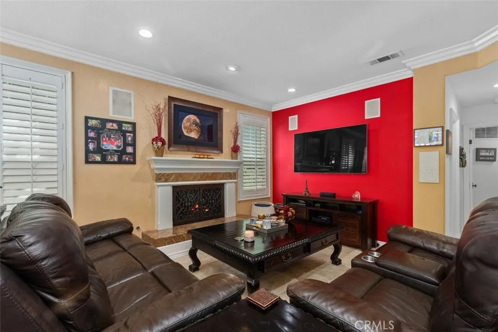 The inviting family room features a classic fireplace with decorative surround, crown molding, recessed lighting, plantation shutters, and a bold accent wall that adds warmth and personality to the space.