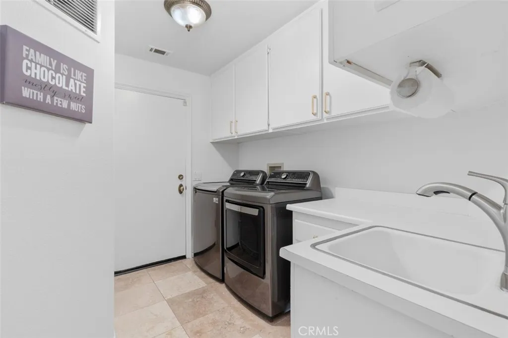 The spacious indoor laundry room features a utility sink, white cabinetry with gold hardware, travertine tile floors, and direct access to the three car garage with epoxy flooring, built in storage cabinets, and Tesla charger.