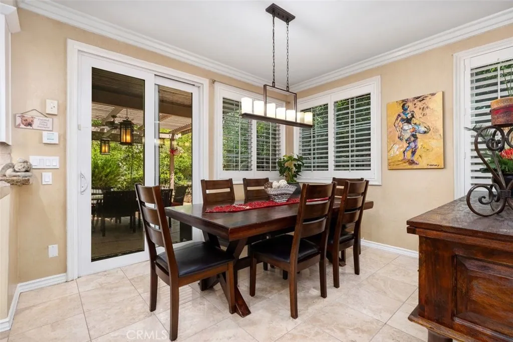 The bright and airy casual dining area features crown molding, travertine tile floors, plantation shutters, and a stylish linear pendant chandelier. French doors open directly to the covered poolside lanai, creating an indoor outdoor flow perfect for everyday dining and entertaining.