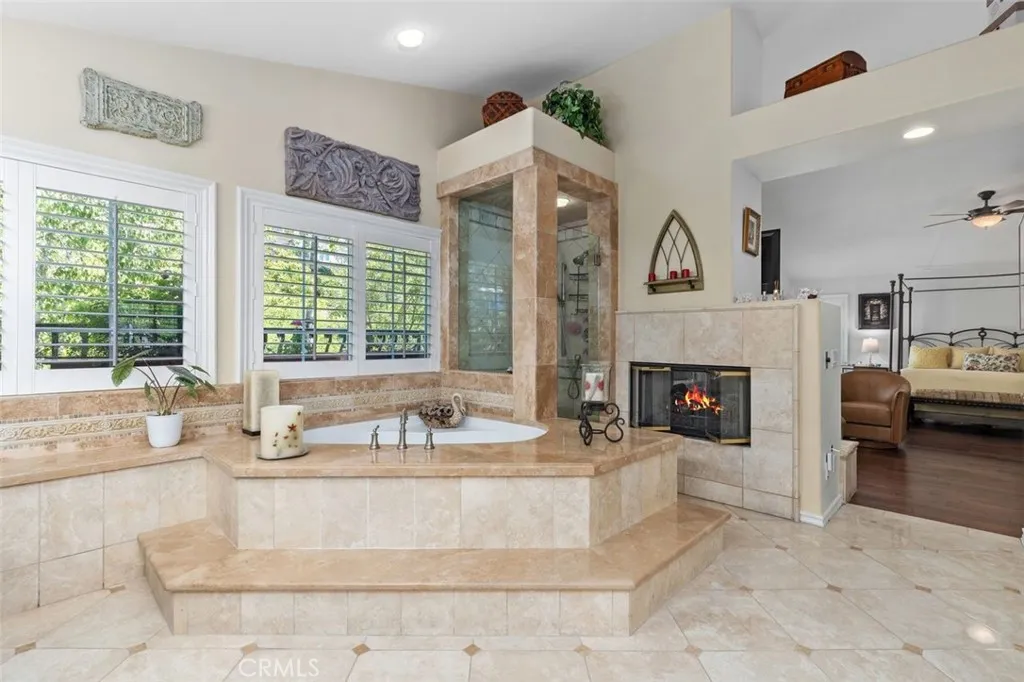 The spa like primary bath features a soaking tub with travertine surround, a glass enclosed shower, and a two sided fireplace shared with the primary suite sitting area. High ceilings, plantation shutters, and elegant tile detail create a luxurious and tranquil retreat.