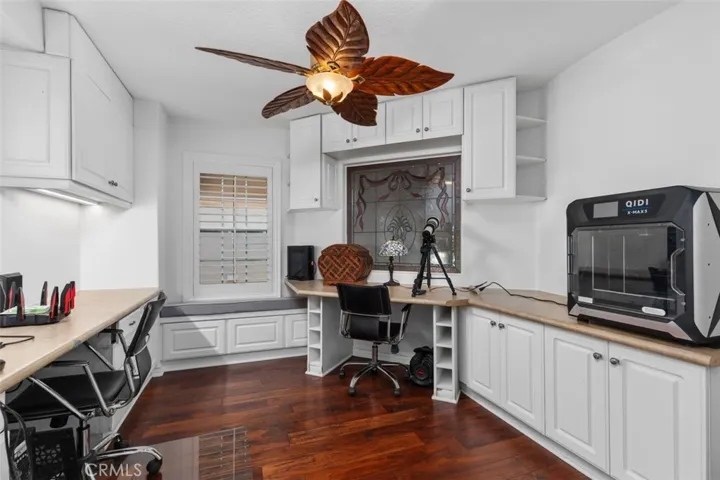 The fifth bedroom is currently configured as a home office featuring wraparound custom built in cabinetry and desk space, rich hardwood floors, plantation shutters, and a unique decorative ceiling fan. A stained glass window accent and abundant storage including under the window seat make this an ideal workspace or creative studio.