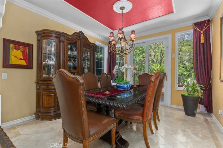 The formal dining room is anchored by a custom tray ceiling with medallion detail, elegant chandelier, and crown molding. French doors and large windows overlook the lush garden and pool beyond, filling the room with natural light.