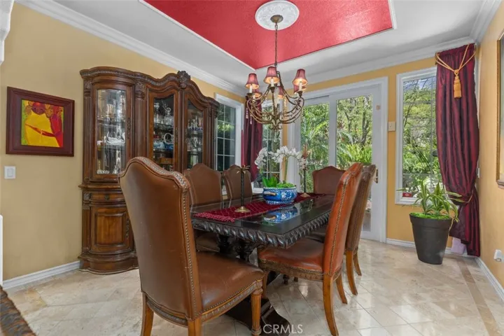 The formal dining room is anchored by a custom tray ceiling with medallion detail, elegant chandelier, and crown molding. French doors and large windows overlook the lush garden and pool beyond, filling the room with natural light.