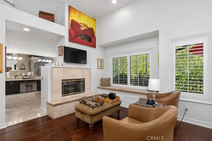 The primary suite sitting area features warm hardwood floors, a two sided fireplace with travertine surround, a built in window seat with storage, plantation shutters, and soaring ceilings with recessed lighting. An open view into the spa bath beyond creates a seamless and luxurious flow between living and retreat spaces.