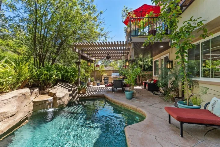 This stunning backyard view showcases the sparkling pool with custom rock waterfall, the covered pergola lanai with ceiling fan and pendant lanterns, the built in BBQ island with beverage fridge, and the private primary suite balcony above. Lush mature tropical landscaping surrounds every corner of this outdoor retreat, creating a truly private and resort style living experience.