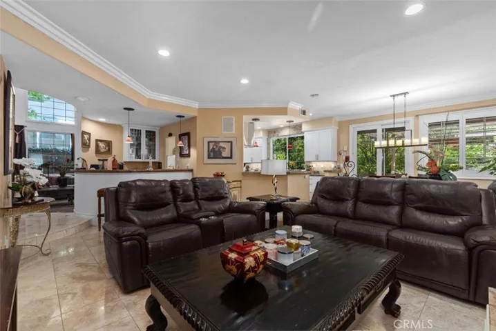 This expansive view captures the seamless open flow between the family room, granite topped bar, remodeled kitchen, and casual dining area all united by travertine tile floors, crown molding, and recessed lighting. Large windows and plantation shutters frame lush garden views on every side, filling the space with natural light throughout the day.