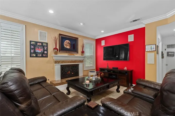 The inviting family room features a classic fireplace with decorative surround, crown molding, recessed lighting, plantation shutters, and a bold accent wall that adds warmth and personality to the space.