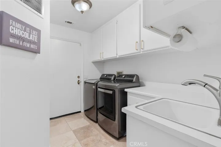 The spacious indoor laundry room features a utility sink, white cabinetry with gold hardware, travertine tile floors, and direct access to the three car garage with epoxy flooring, built in storage cabinets, and Tesla charger.