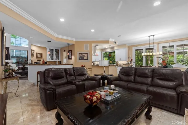 This expansive view captures the seamless open flow between the family room, granite topped bar, remodeled kitchen, and casual dining area all united by travertine tile floors, crown molding, and recessed lighting. Large windows and plantation shutters frame lush garden views on every side, filling the space with natural light throughout the day.