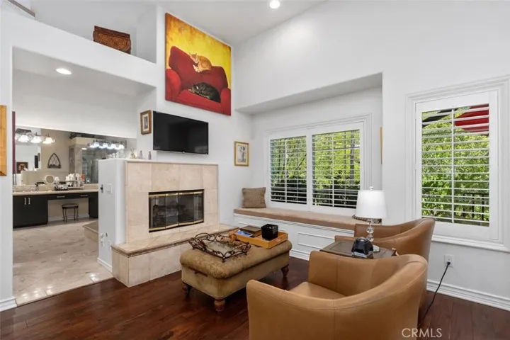 The primary suite sitting area features warm hardwood floors, a two sided fireplace with travertine surround, a built in window seat with storage, plantation shutters, and soaring ceilings with recessed lighting. An open view into the spa bath beyond creates a seamless and luxurious flow between living and retreat spaces.