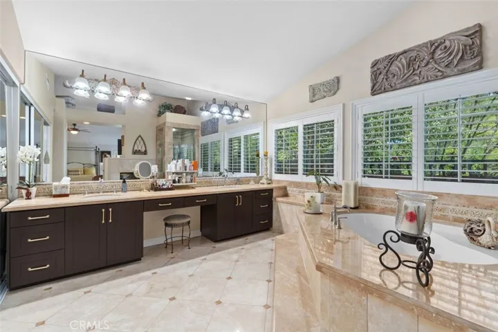 The expansive primary bathroom features a generous dual sink travertine vanity with dark cabinetry and gold hardware, and a wall to wall mirror with Hollywood style lighting above. Plantation shutters frame lush treetop views while the soaking tub and glass enclosed shower are visible beyond, completing this serene and luxurious spa retreat.