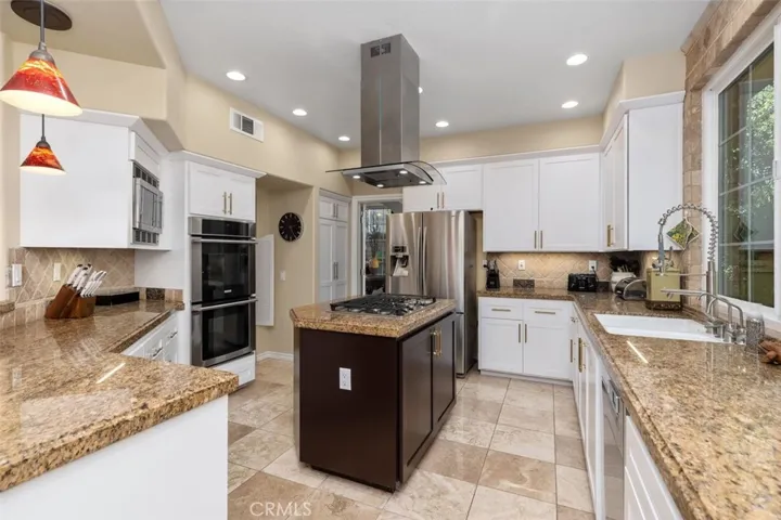The remodeled chef's kitchen features granite countertops, stainless steel appliances, and a 5-burner gas cooktop island with a dramatic stainless range hood. White shaker cabinetry with brushed gold hardware and a travertine tile backsplash complete this stunning and functional space.