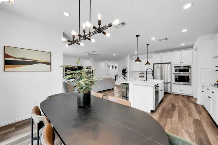 Open-concept living space featuring wood-finish flooring, a contemporary dining area with a linear chandelier, a kitchen island with pendant lighting and bar seating, white cabinetry, and stainless steel appliances