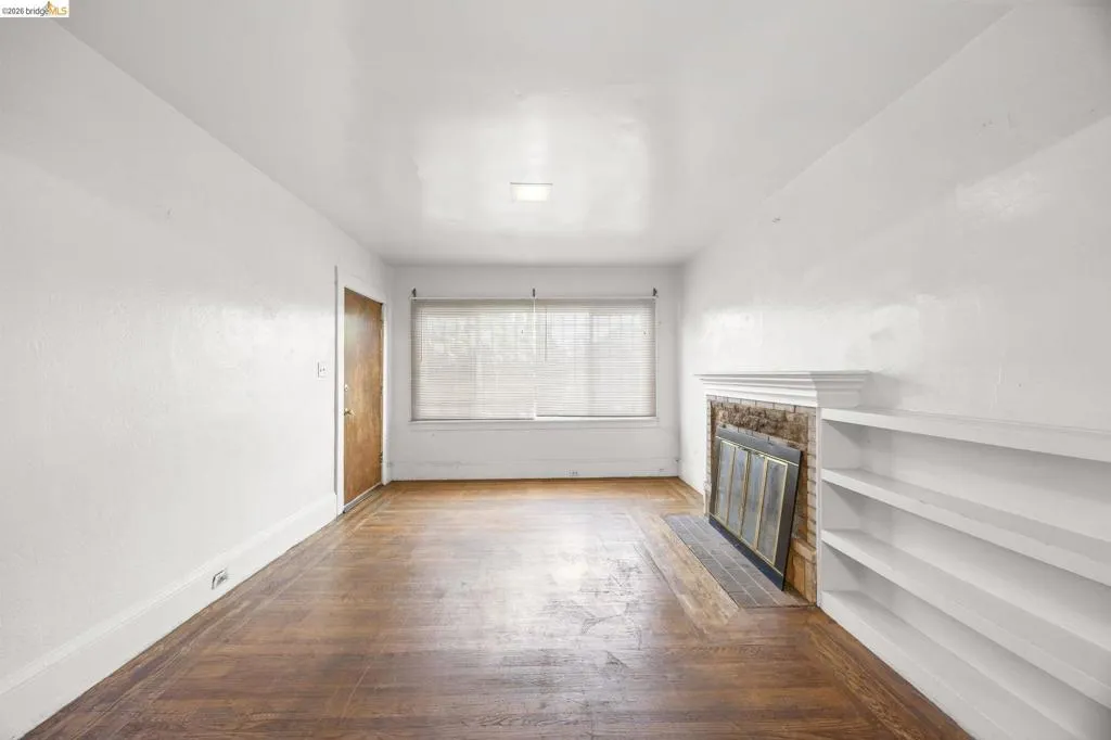 Living space featuring wood-finish flooring, a brick fireplace with a white mantel, built-in shelving, a window with blinds, and white painted walls