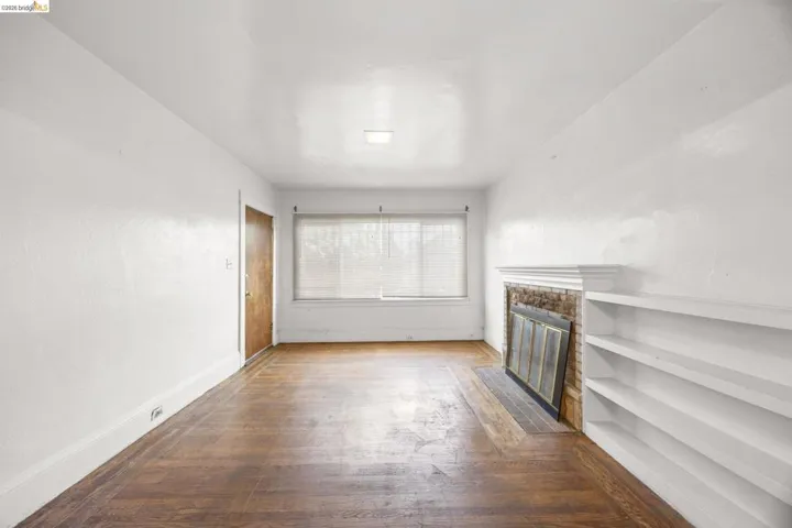 Living space featuring wood-finish flooring, a brick fireplace with a white mantel, built-in shelving, a window with blinds, and white painted walls