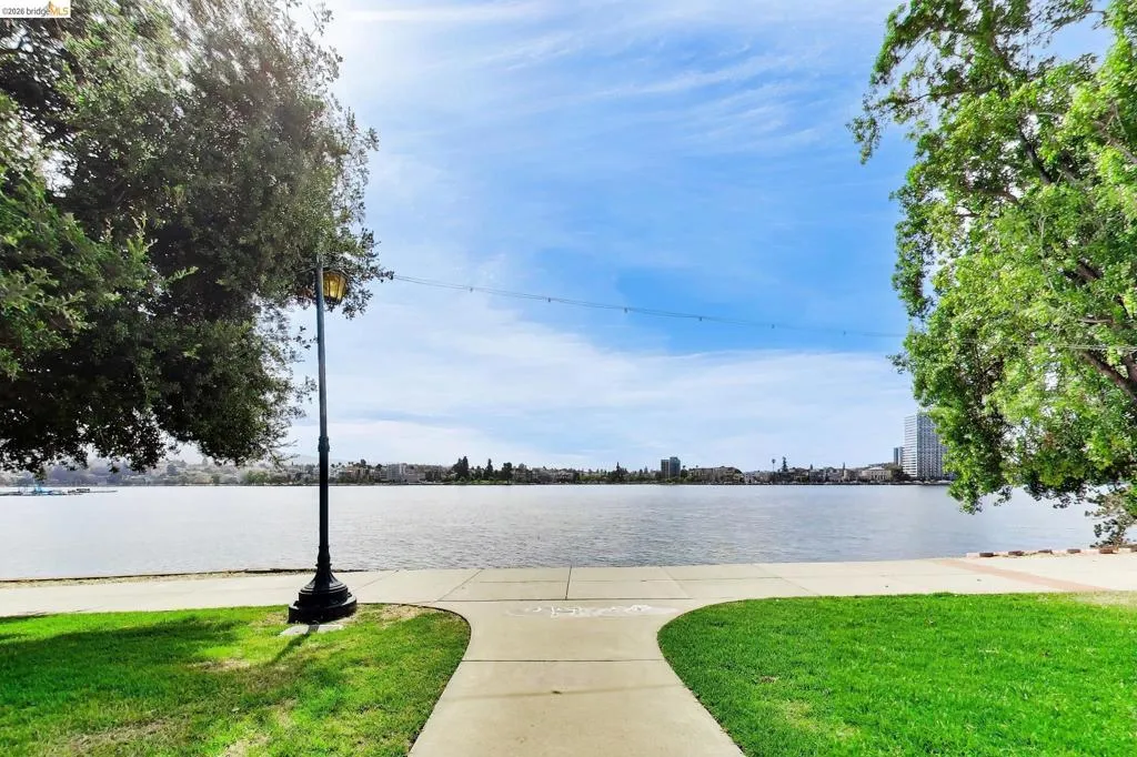 Waterfront park pathway with a classic streetlamp, mature trees, and a distant cityscape across the water