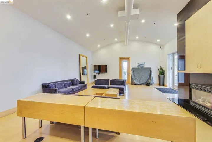 Spacious open-concept room featuring a vaulted ceiling with recessed lighting, light wood-finish flooring, and a built-in fireplace with a dark stone surround and light wood cabinetry