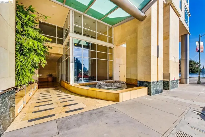 Modern building entrance featuring a glass-enclosed lobby, a built-in water feature, natural stone cladding, a covered walkway, and mature landscaping