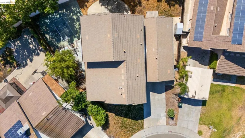 Aerial perspective showcasing a property with a shingle roof, concrete driveway, and a paved sport court in the backyard