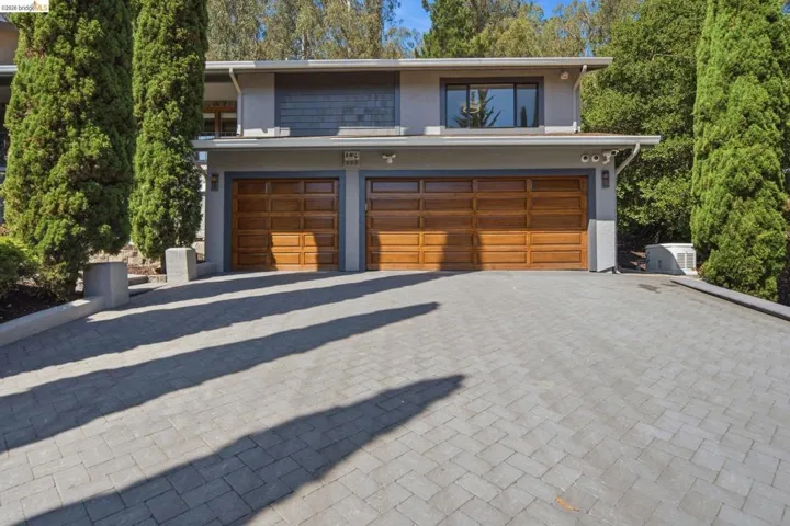 Contemporary exterior with a two-car garage featuring wood-paneled doors, a paver driveway, and mature vertical landscaping