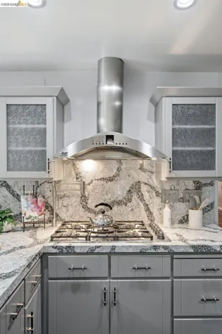 Kitchen featuring a stainless steel range hood, built-in gas cooktop, patterned countertop and backsplash, and light gray cabinetry with dark hardware