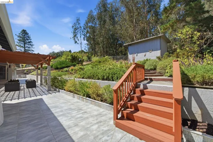 Expansive outdoor patio featuring large format tile, a wood pergola with stone-wrapped columns, and a terraced hillside with mature landscaping