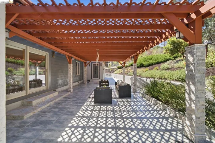 Expansive outdoor patio featuring a wooden pergola, patterned tile flooring, sliding glass doors, stone-clad support columns, and built-in exterior lighting